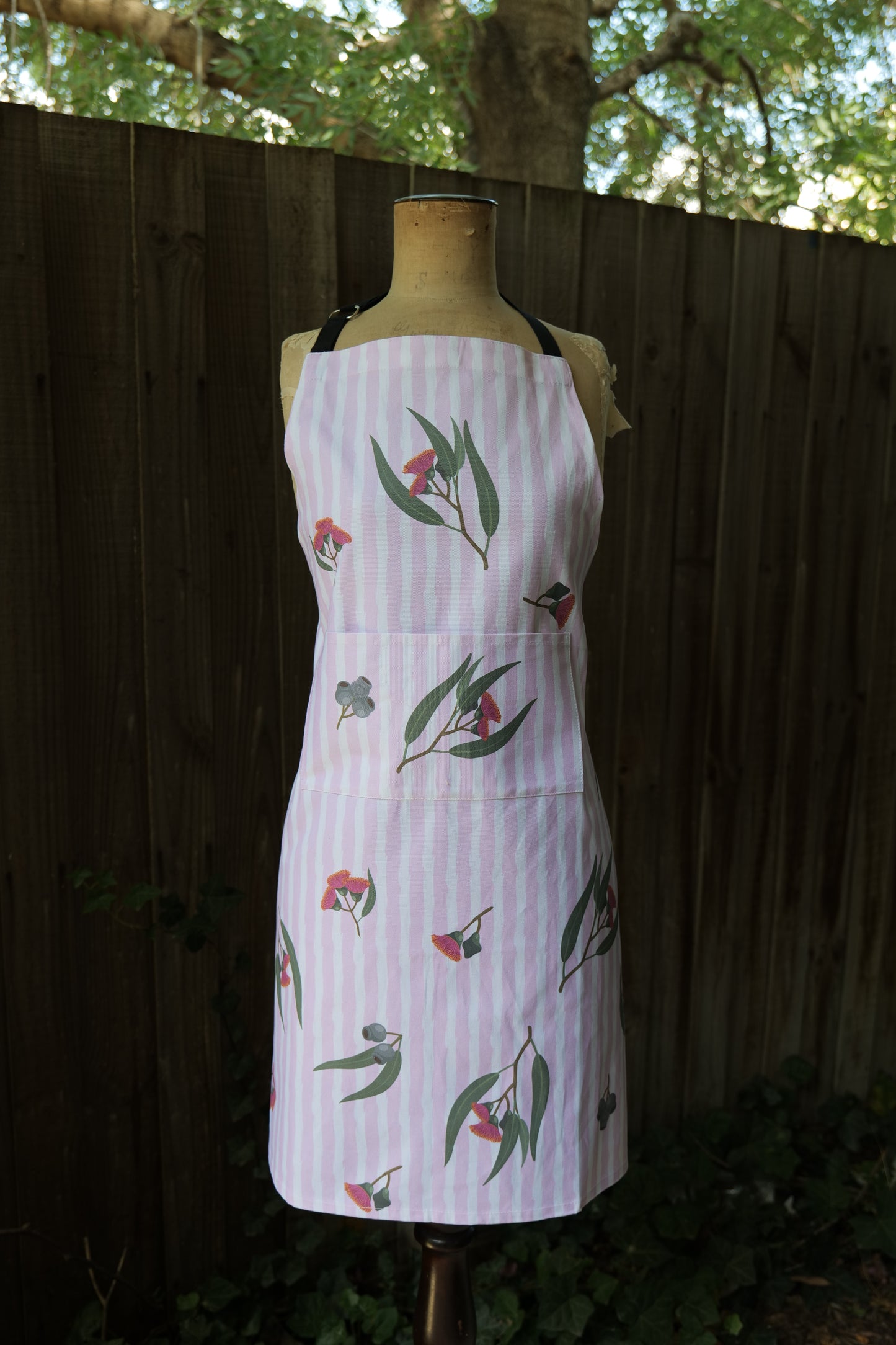 Apron in Eucalyptus design, with pink and white vertical stripes featuring illustrated eucalyptus leaves and blossoms; apron has an adjustable neck strap, extended waist ties and front pocket; generous fit, handmade in Australia from cotton canvas and machine washable, modelled on a mannequin in front of a dark background