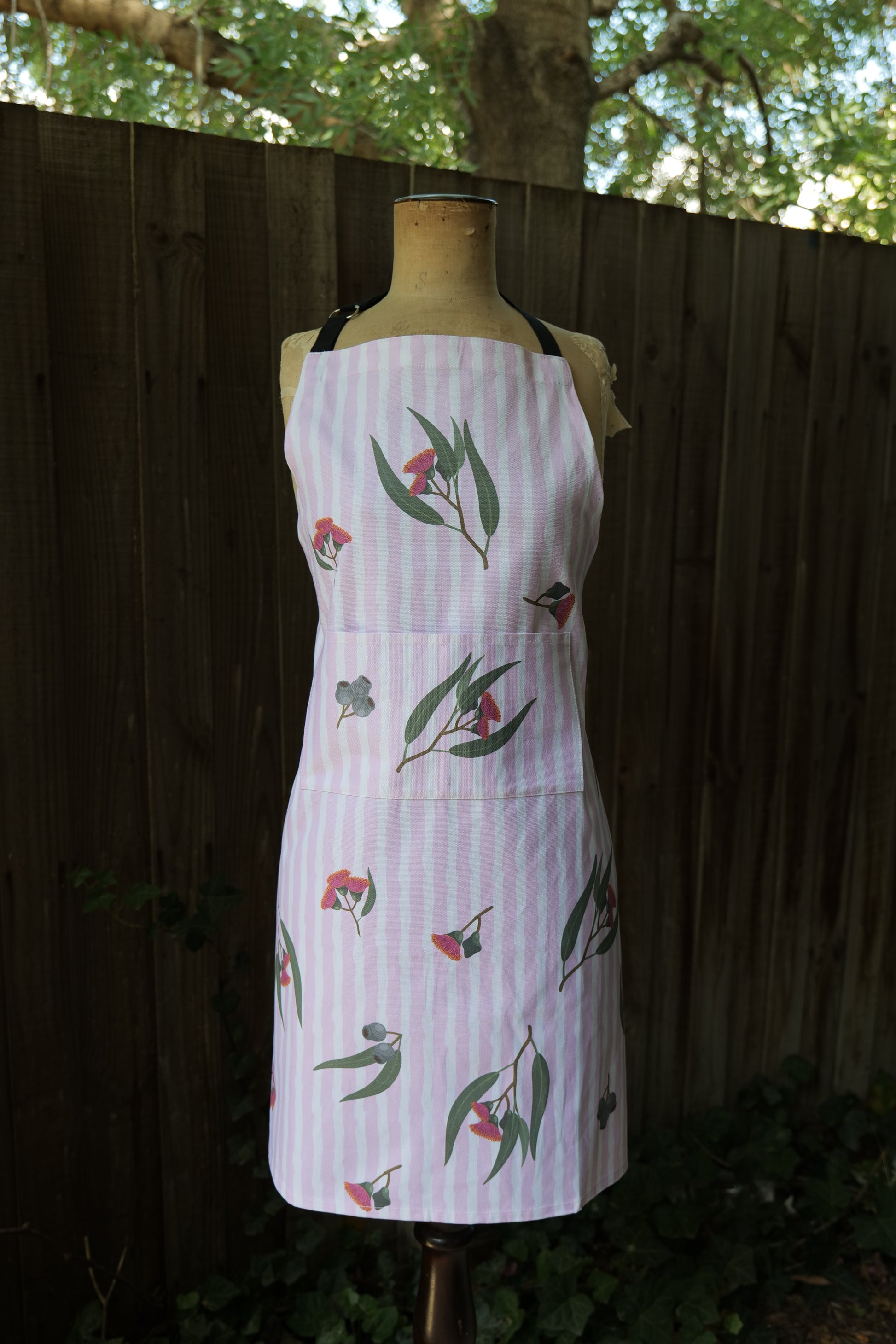 Apron in Eucalyptus design, with pink and white vertical stripes featuring illustrated eucalyptus leaves and blossoms; apron has an adjustable neck strap, extended waist ties and front pocket; generous fit, handmade in Australia from cotton canvas and machine washable, modelled on a mannequin in front of a dark background