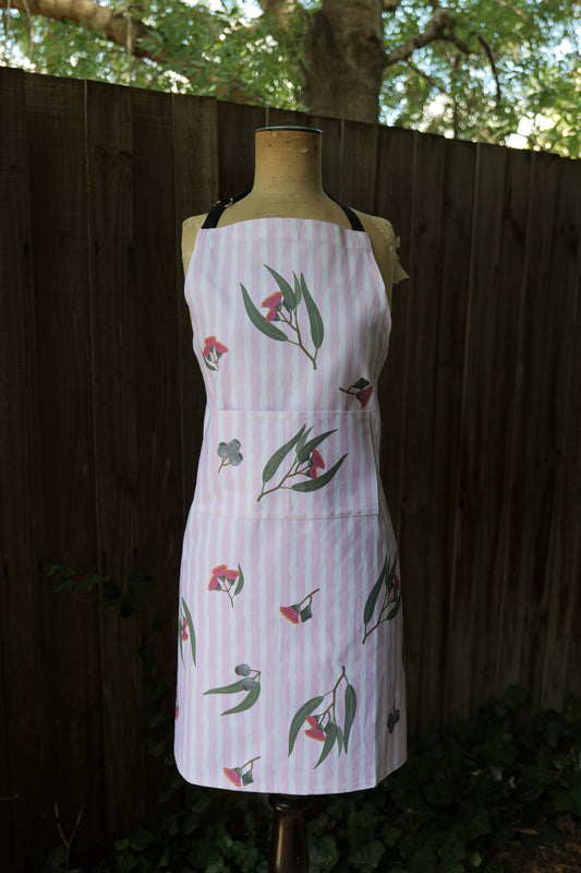Apron in Eucalyptus design, with pink and white vertical stripes featuring illustrated eucalyptus leaves and blossoms; apron has an adjustable neck strap, extended waist ties and front pocket; generous fit, handmade in Australia from cotton canvas and machine washable, modelled on a mannequin in front of a dark background