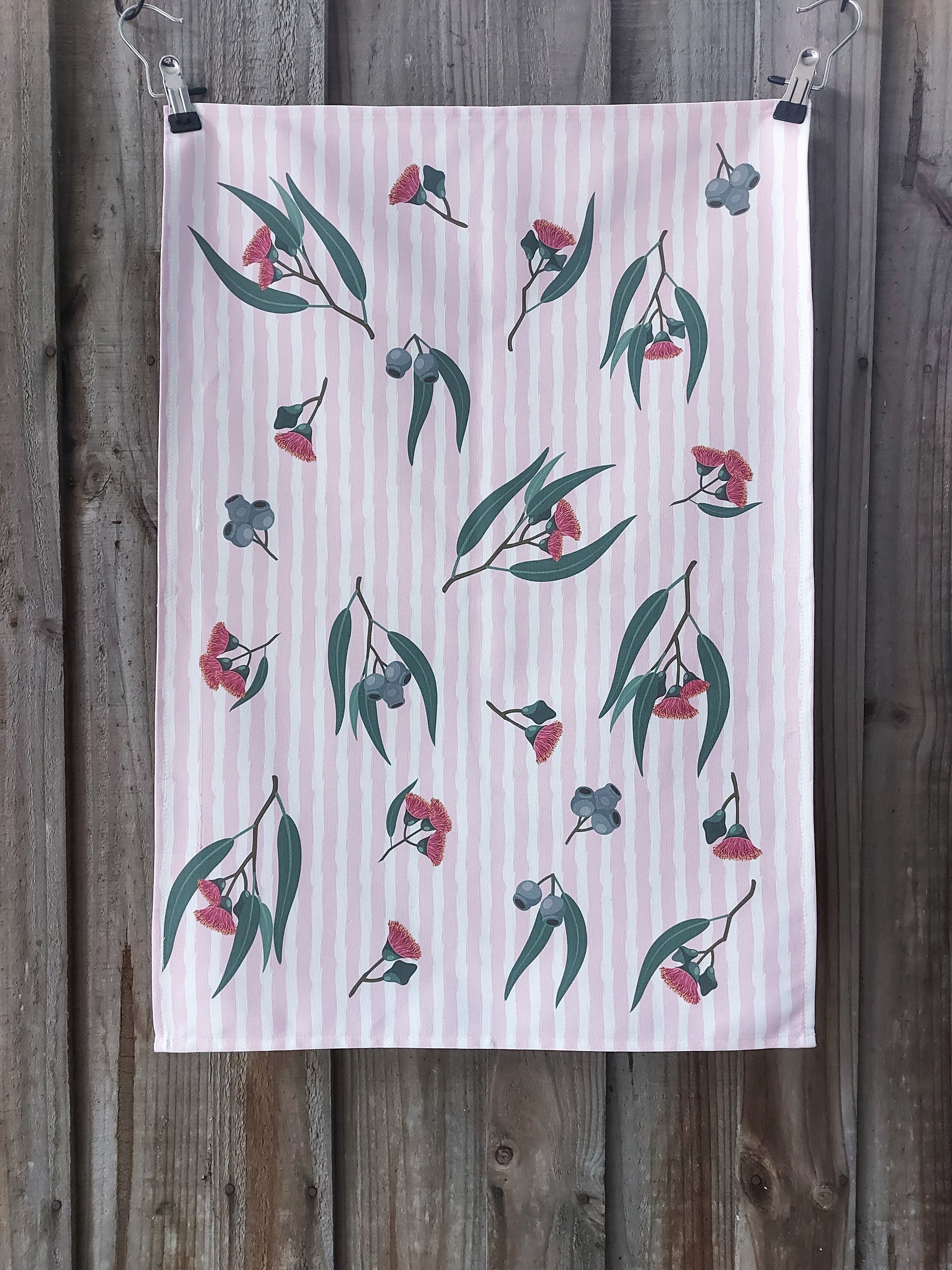 Tea towel featuring pink eucalyptus blossom and gum nuts on a pink and white striped background, displayed against a wooden fence