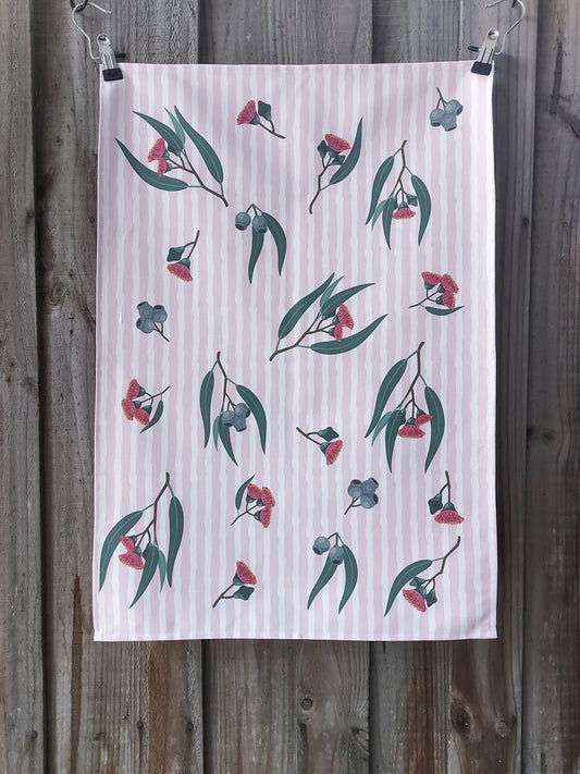 Tea towel featuring pink eucalyptus blossom and gum nuts on a pink and white striped background, displayed against a wooden fence