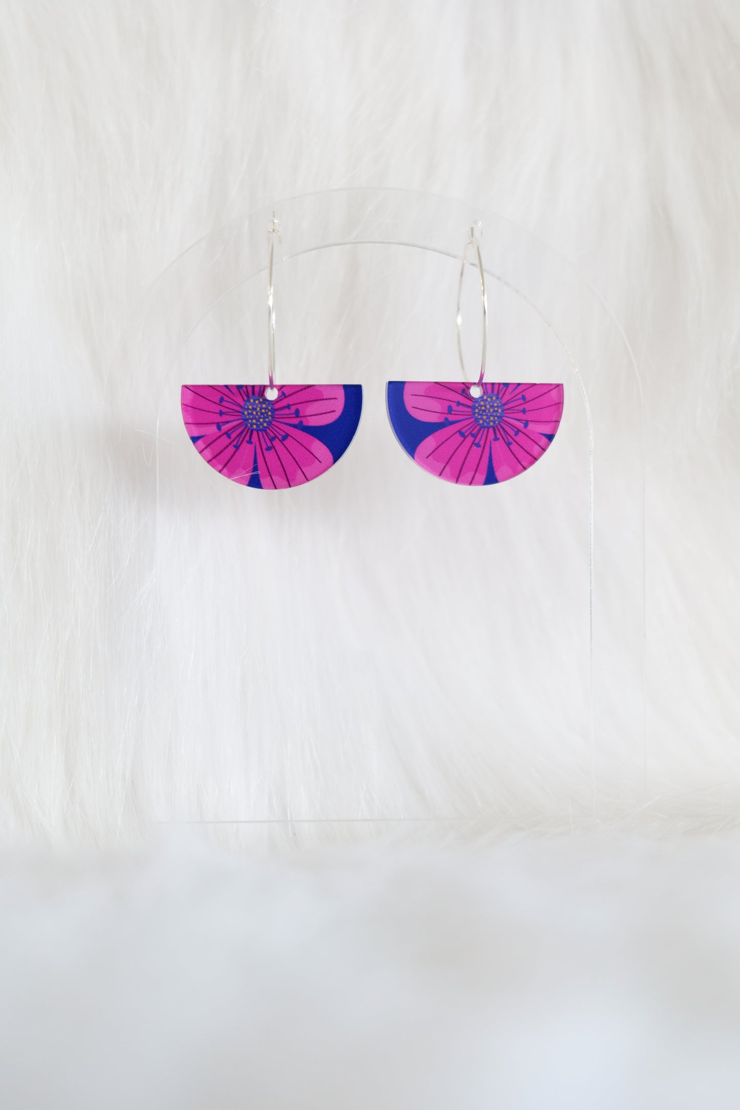 Half-moon shaped earrings featuring a bright pink flower on a dark blue background, hanging from silver hoops, displayed on a clear stand against a white background