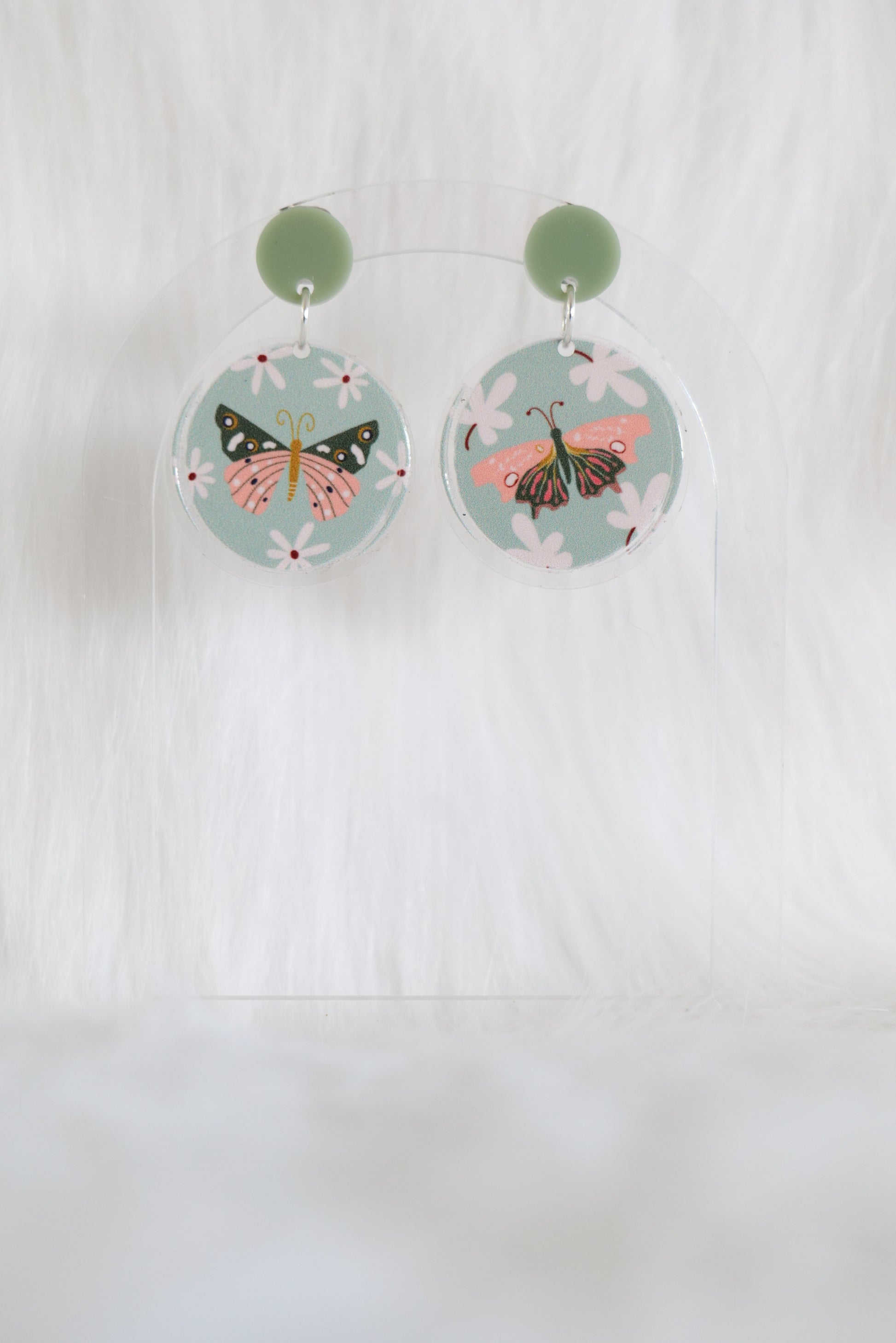 Pair of mismatched round earrings featuring pink and olive green butterflies on a pale green background with cream flowers, printed on lightweight acrylic with stainless steel findings and large backs; designed and made in Australia