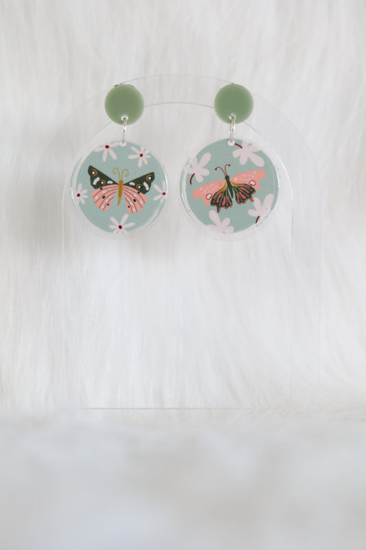 Pair of mismatched round earrings featuring pink and olive green butterflies on a pale green background with cream flowers, printed on lightweight acrylic with stainless steel findings and large backs; designed and made in Australia