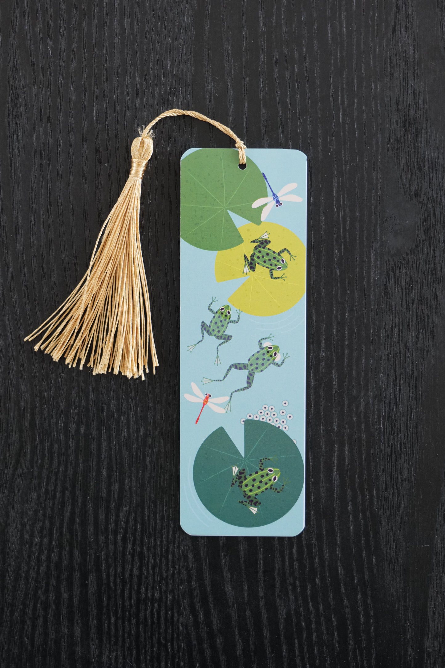 Bookmark with frogs and lily pads on a blue background and a gold tassel, sitting against a black backdrop