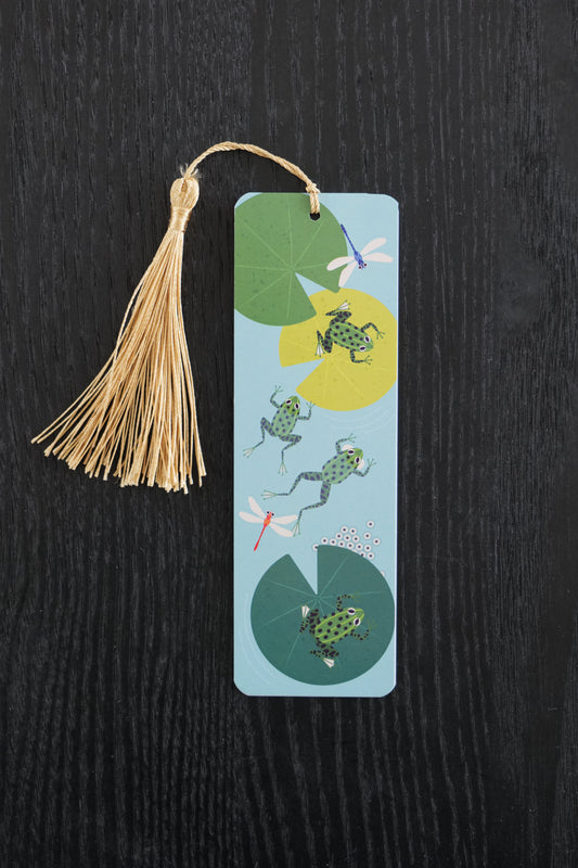 Bookmark with frogs and lily pads on a blue background and a gold tassel, sitting against a black backdrop