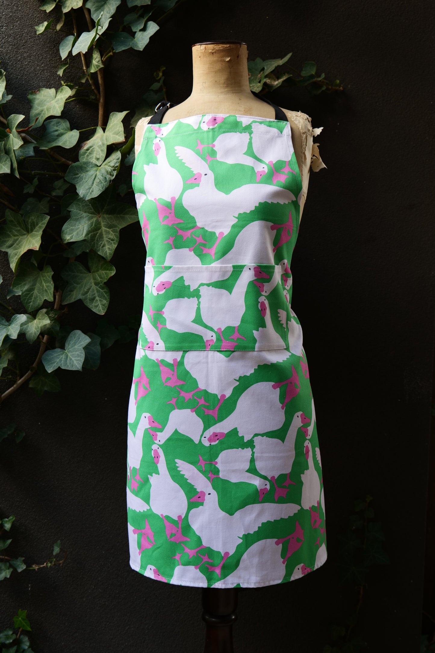 Bright green apron featuring playful white geese with hot pink beaks and feet, on a mannequin in front of a dark background, apron is generously sized with adjustable neck and long waist ties, front pocket visible. Made from premium heavyweight cotton, Australian made.