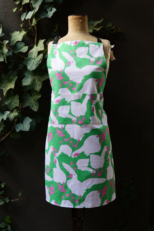 Bright green apron featuring playful white geese with hot pink beaks and feet, on a mannequin in front of a dark background, apron is generously sized with adjustable neck and long waist ties, front pocket visible. Made from premium heavyweight cotton, Australian made.