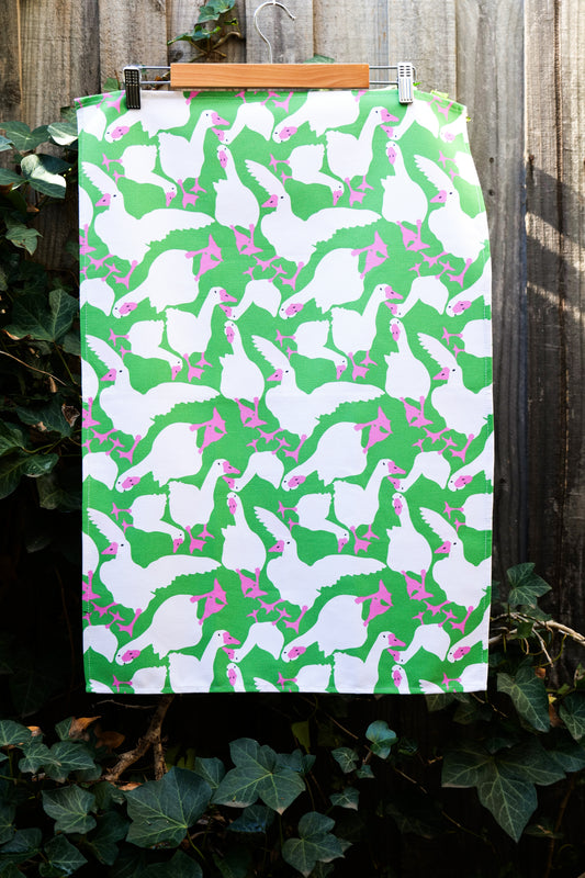 Tea towel featuring lots of white geese in various positions with hot pink beaks and feet on a bright green background; tea towel displayed against a wooden fence