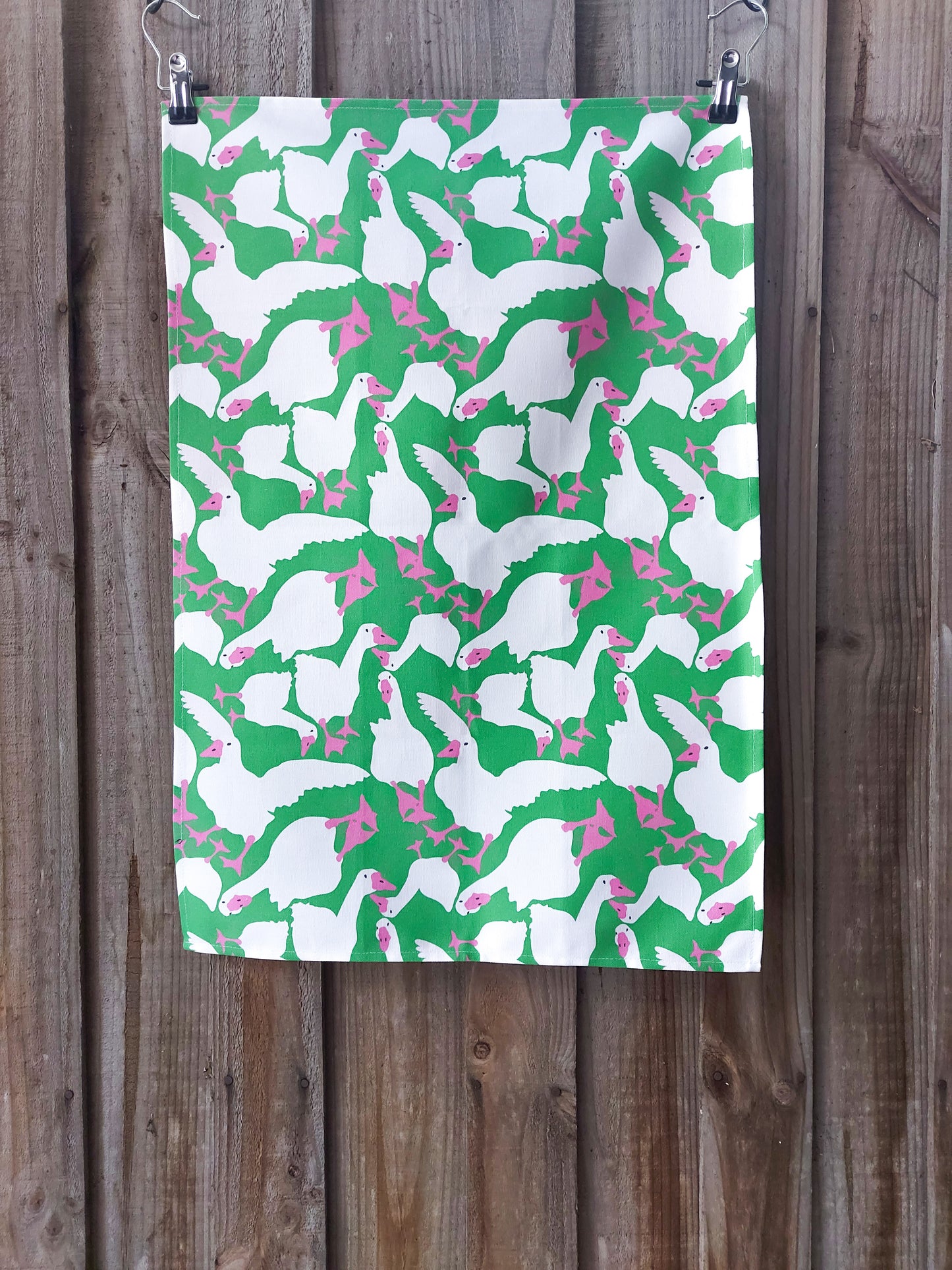 Tea towel featuring white geese in various poses with pink beaks and feet against a bright green background; displayed against a wooden fence