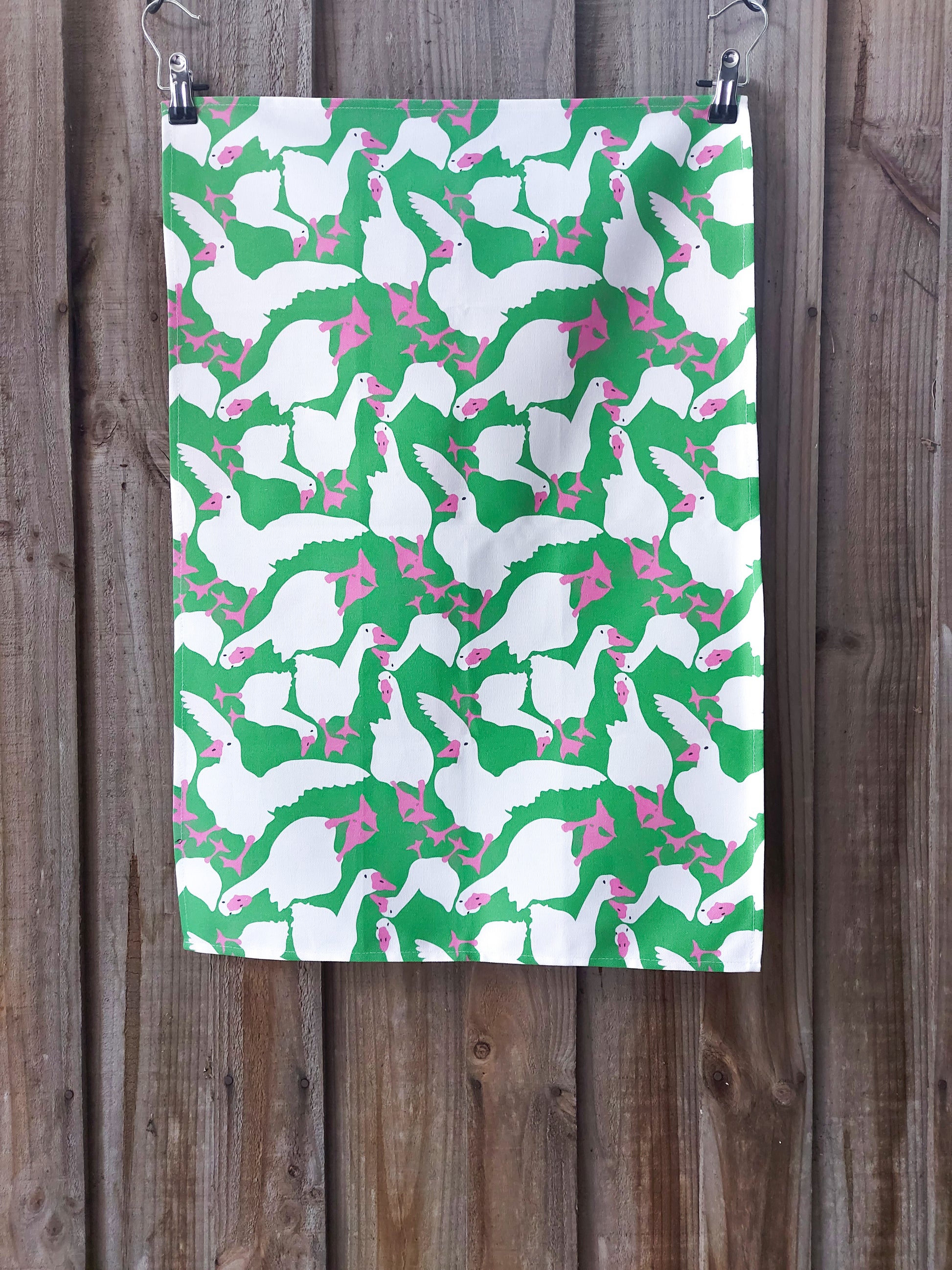 Tea towel featuring white geese in various poses with pink beaks and feet against a bright green background; displayed against a wooden fence