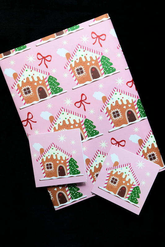Sheet of folded gift wrap featuring gingerbread houses, stars and red bows on a pink background with two matching gift cards, displayed against a black backdrop