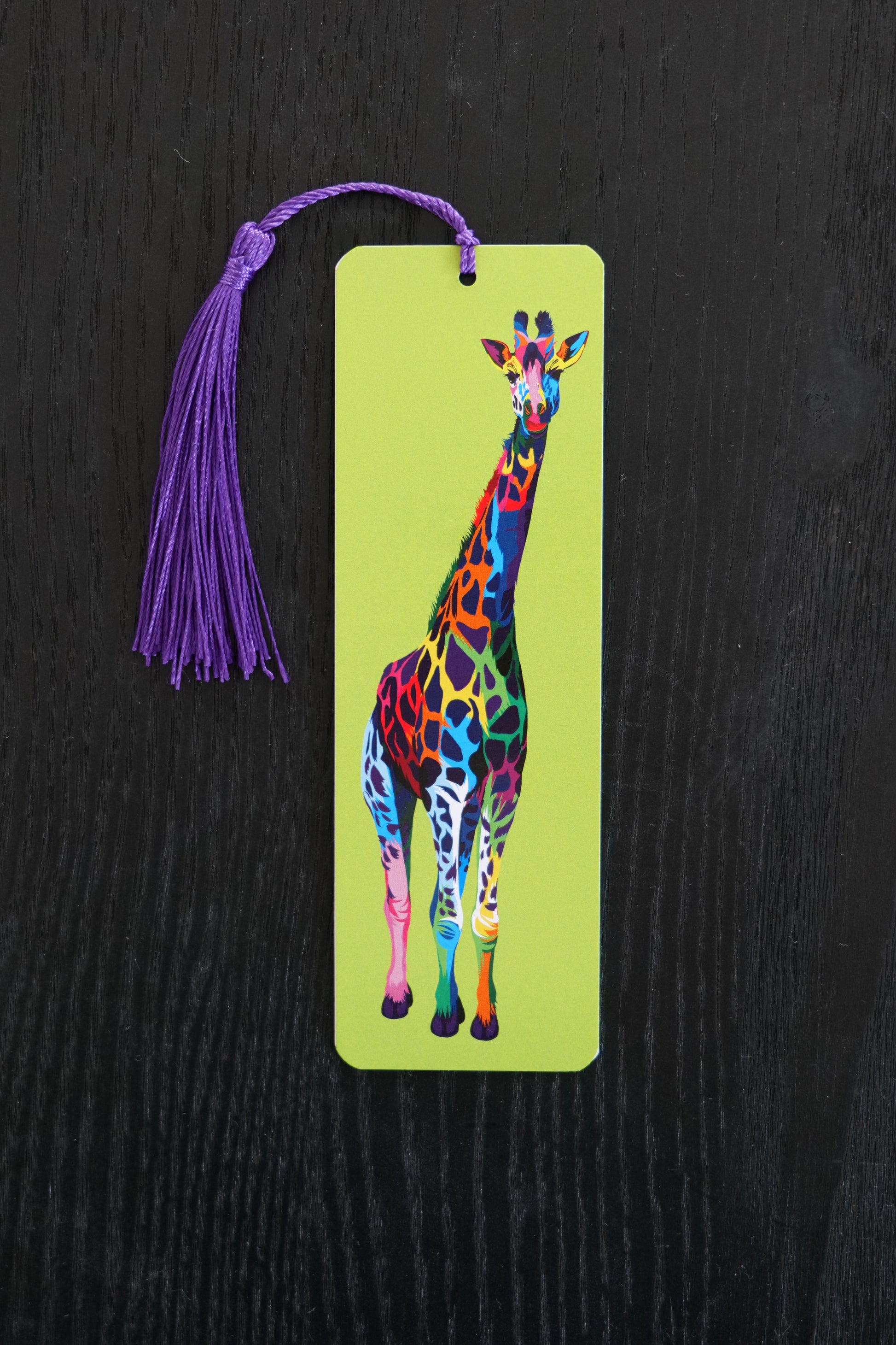 Bookmark with a multicoloured giraffe against a lime green background, with a purple tassel; sitting against a black backdrop