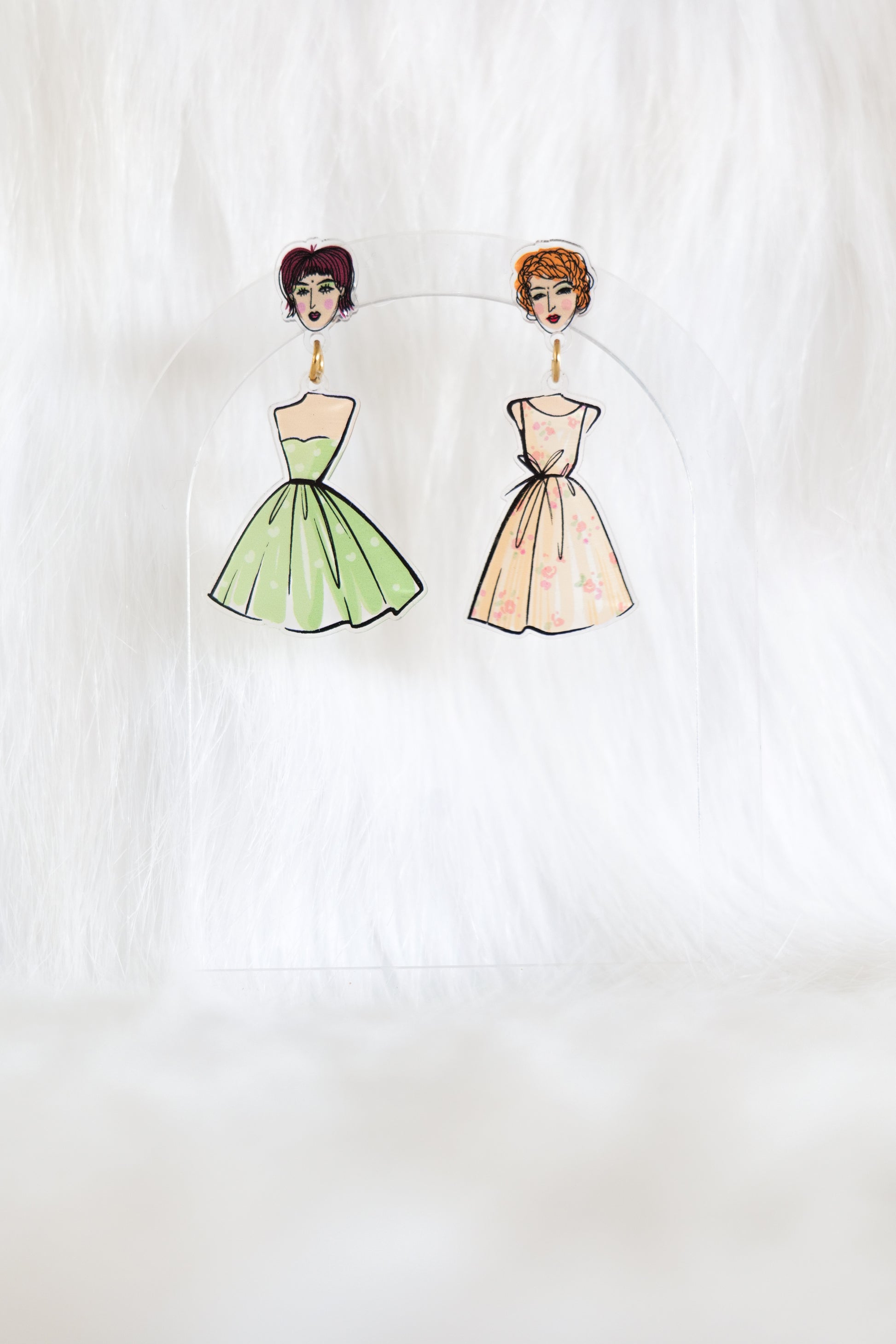 Mismatched pair of earrings with girls faces as the stud tops, one has a green spotty dress hanging beneath and the other has a peach floral dress; displayed on a clear stand against a white background