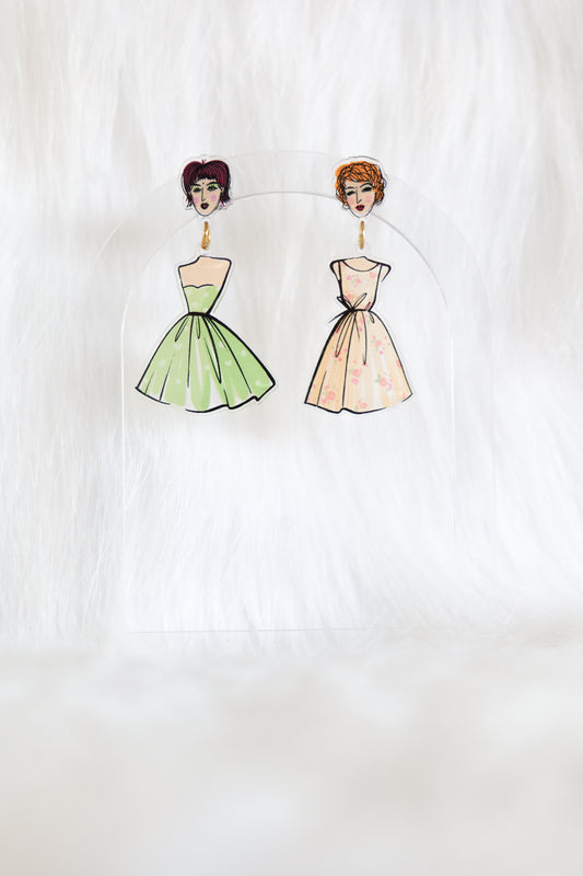 Mismatched pair of earrings with girls faces as the stud tops, one has a green spotty dress hanging beneath and the other has a peach floral dress; displayed on a clear stand against a white background