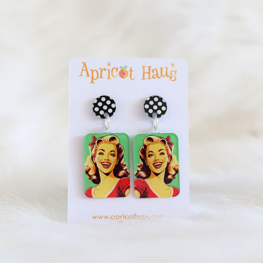Pair of rectangular earrings featuring a pop-art style smiling woman on a bright green background, with a circular black and white spotted stud top, printed on lightweight acrylic with stainless steel post and large accessible back, displayed on an Apricot Haus earring card