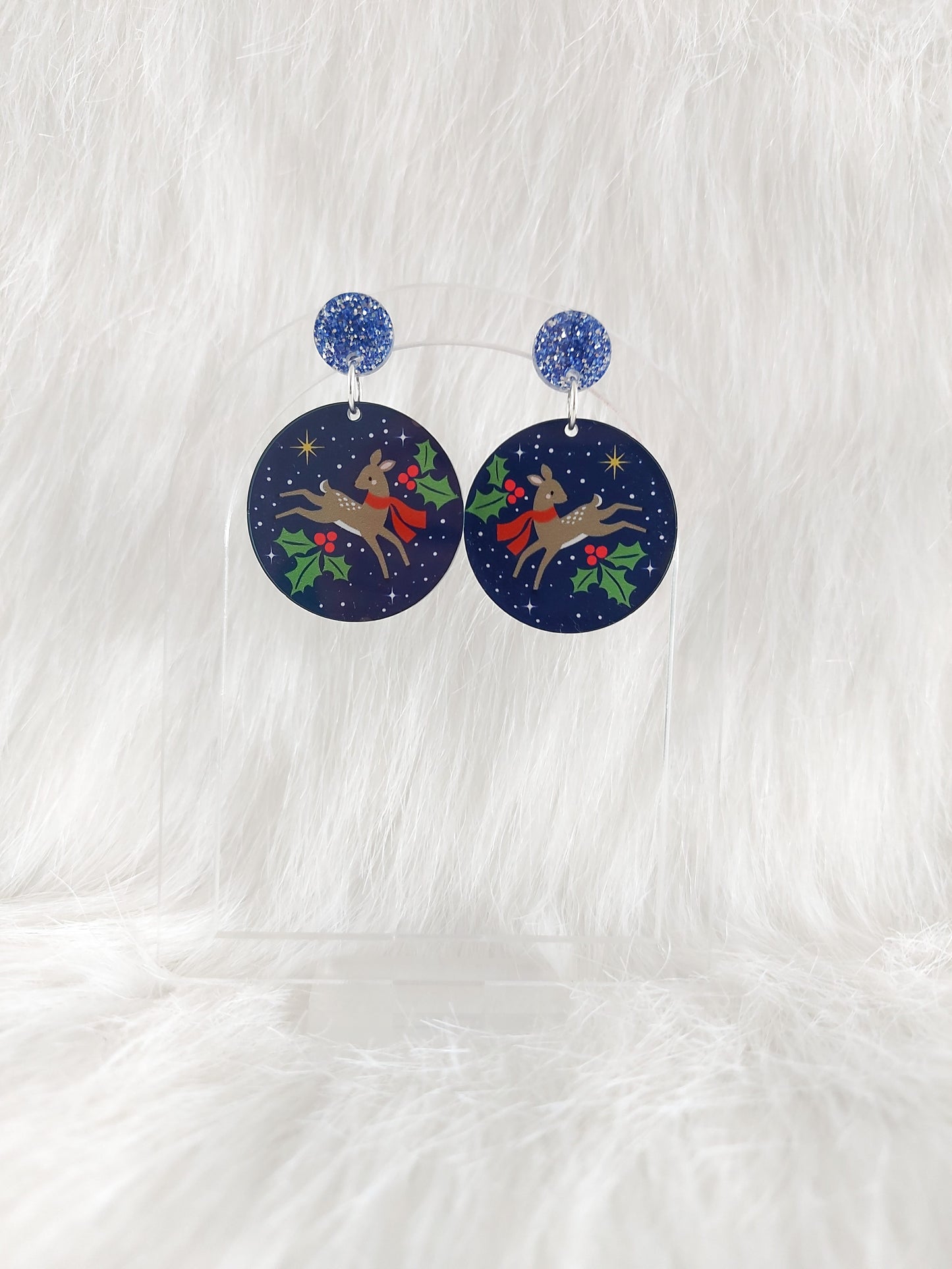 A pair of round navy acrylic earrings featuring a reindeer in a red scarf surrounded by holly and stars, displayed on a clear stand against a white background