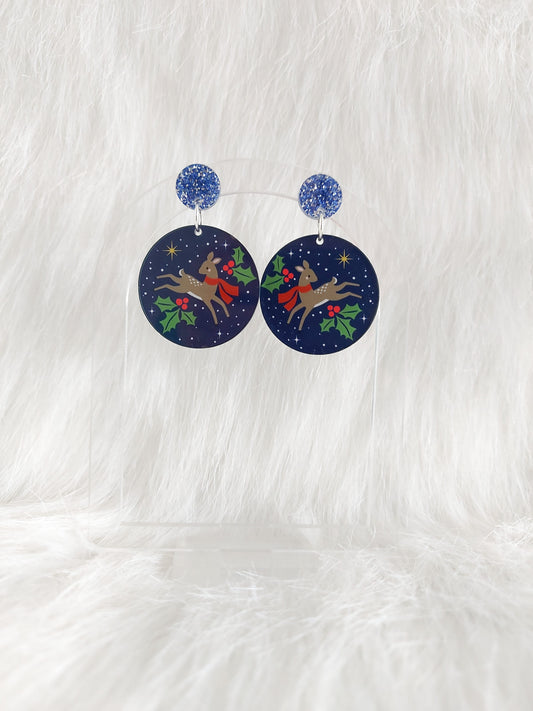 A pair of round navy acrylic earrings featuring a reindeer in a red scarf surrounded by holly and stars, displayed on a clear stand against a white background