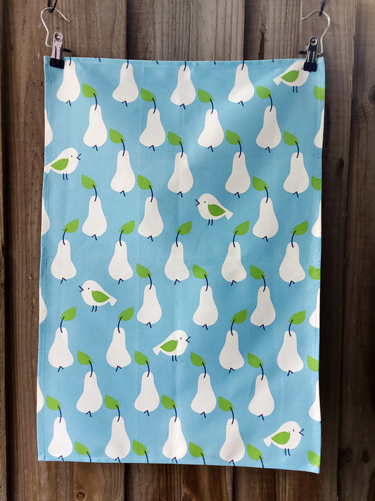 Cotton tea towel featuring a Scandi‑inspired pattern of white pears with green leaves and small birds on a blue background; displayed against a wooden fence