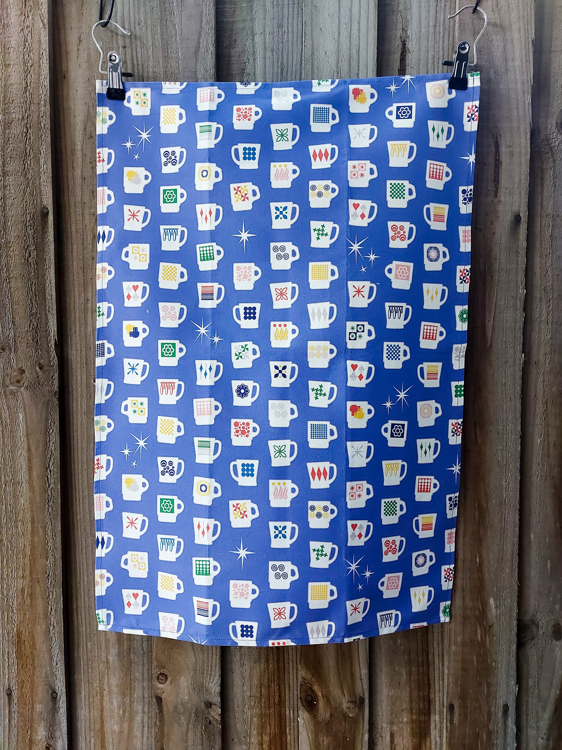 Cotton tea towel featuring lots of white cups, each with a retro or mid-century motif, and some white stars, on a blue/purple background; displayed against a wooden fence