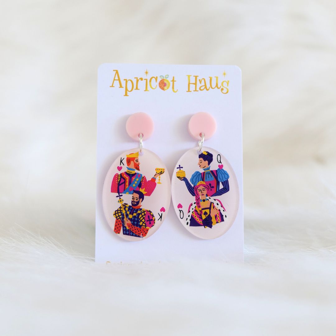 Pair of oval shaped lightweight acrylic earrings featuring two kings on one and two queens on the other, with a pale pink background and top, earrings have stainless steel posts and large accessible backs; made in Australia, displayed on an Apricot Haus earring card