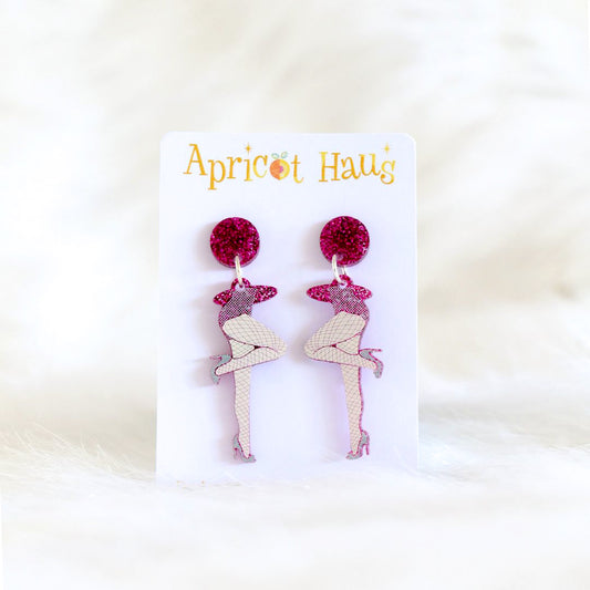 Pair of earrings in the shape of legs wearing stockings and high heels, printed on hot pink glitter acrylic with a pink glitter circular stud, lightweight with stainless steel posts and large backs, displayed on an Apricot Haus earring card
