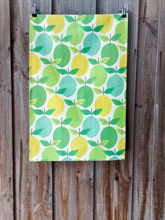 Cotton tea towel with a mid‑century inspired repeating pattern of lemons and limes in yellow, green and teal on a white background; pictured against a wooden fence.