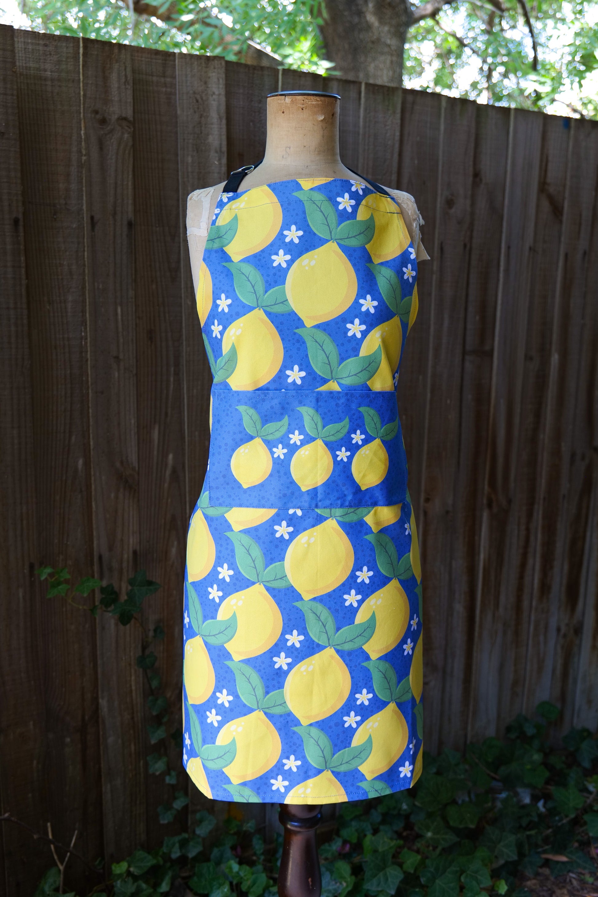Dark blue cotton apron featuring bright yellow lemons with green leaves and small white flowers, displayed on a mannequin in front of a wooden fence