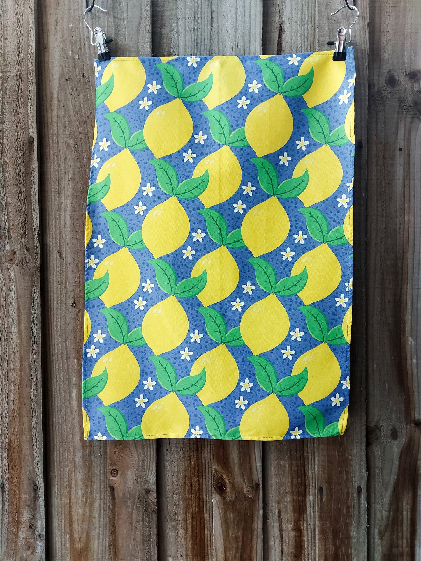 Cotton tea towel featuring bright yellow lemons with green leaves and white blossoms on a blue background; displayed against a wooden fence