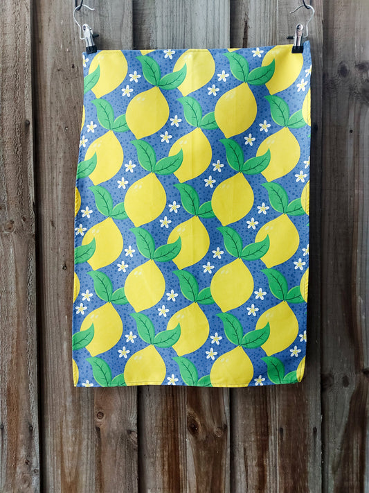 Cotton tea towel featuring bright yellow lemons with green leaves and white blossoms on a blue background; displayed against a wooden fence