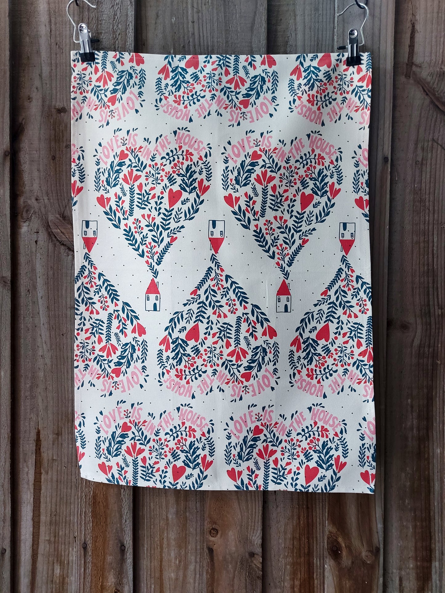 Cotton tea towel featuring Love is in the House design, small nordic inspired houses underneath balloons made of red and navy flowers and words, on a white background; displayed against a wooden fence