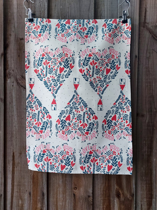 Cotton tea towel featuring Love is in the House design, small nordic inspired houses underneath balloons made of red and navy flowers and words, on a white background; displayed against a wooden fence