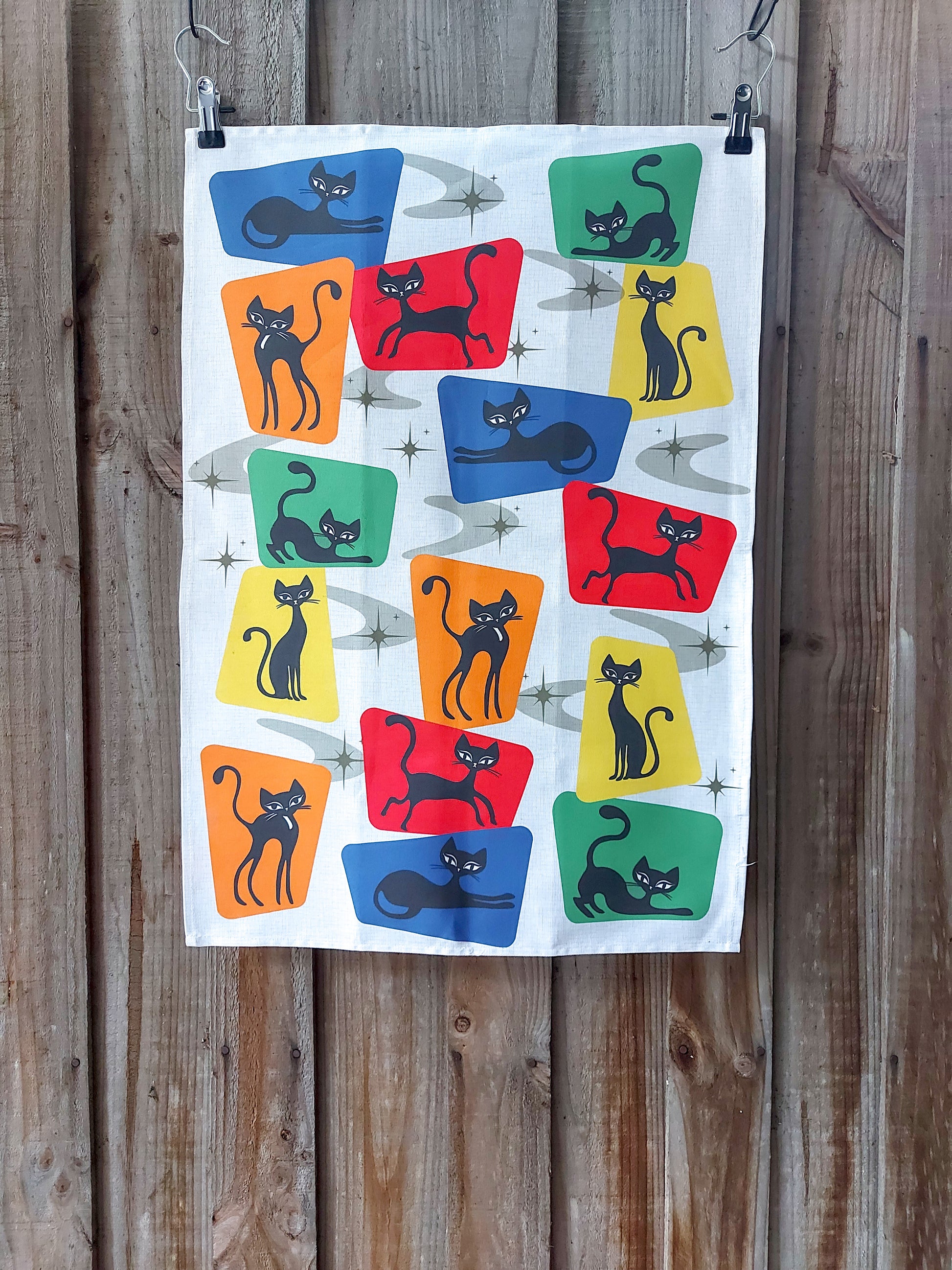 Cotton tea towel featuring 1950s style black cats on brightly coloured blocks on a beige background; displayed against a wooden fence