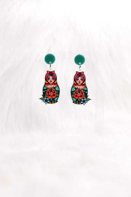 Earrings featuring colourful retro tattoo-style matryoshka dolls with a red rose and a teal stud top, displayed on a clear stand against a white background