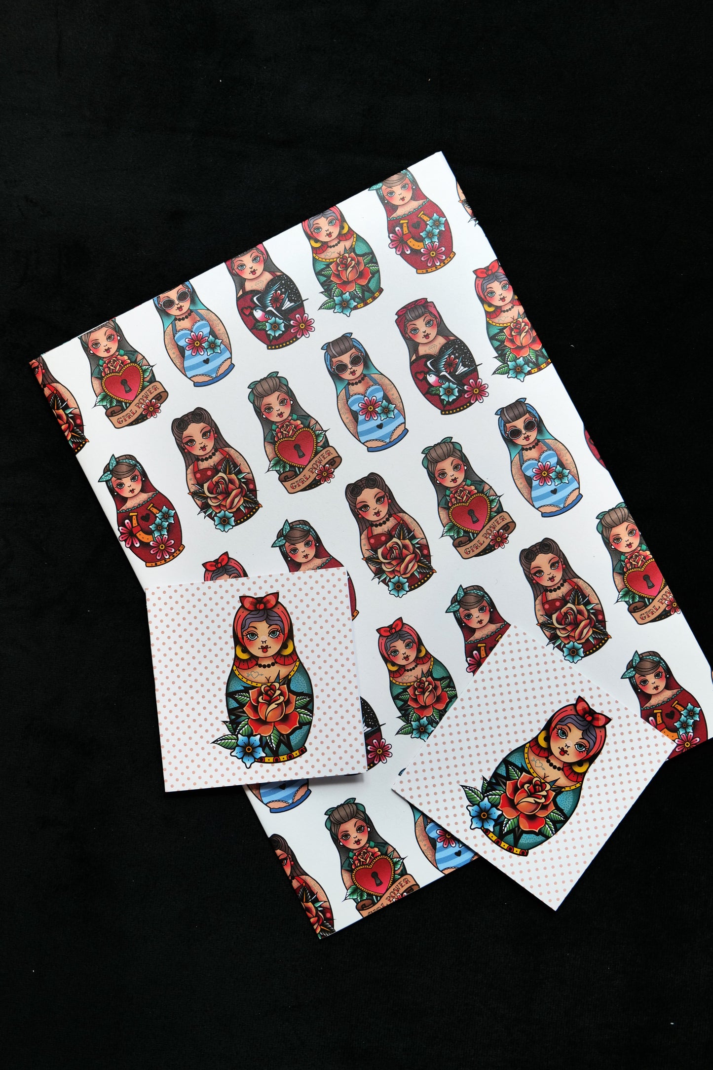 A folded sheet of gift wrap with two matching cards featuring several different retro tattoo-style Matryoshka dolls on a cream background; displayed against a black backdrop