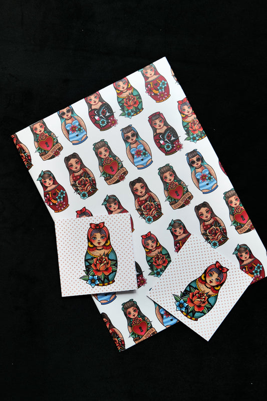A folded sheet of gift wrap with two matching cards featuring several different retro tattoo-style Matryoshka dolls on a cream background; displayed against a black backdrop
