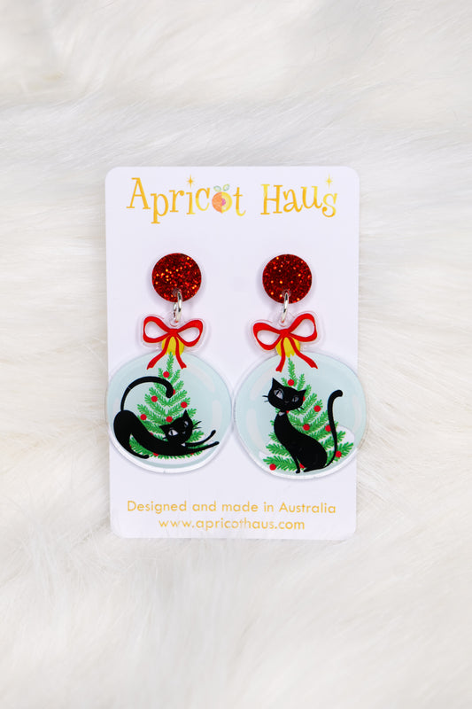 Christmas earrings in the shape of a Christmas bauble with retro-styled black cats and trees, topped with red bows and a red glitter stud; displayed on an Apricot Haus earring card on a white background