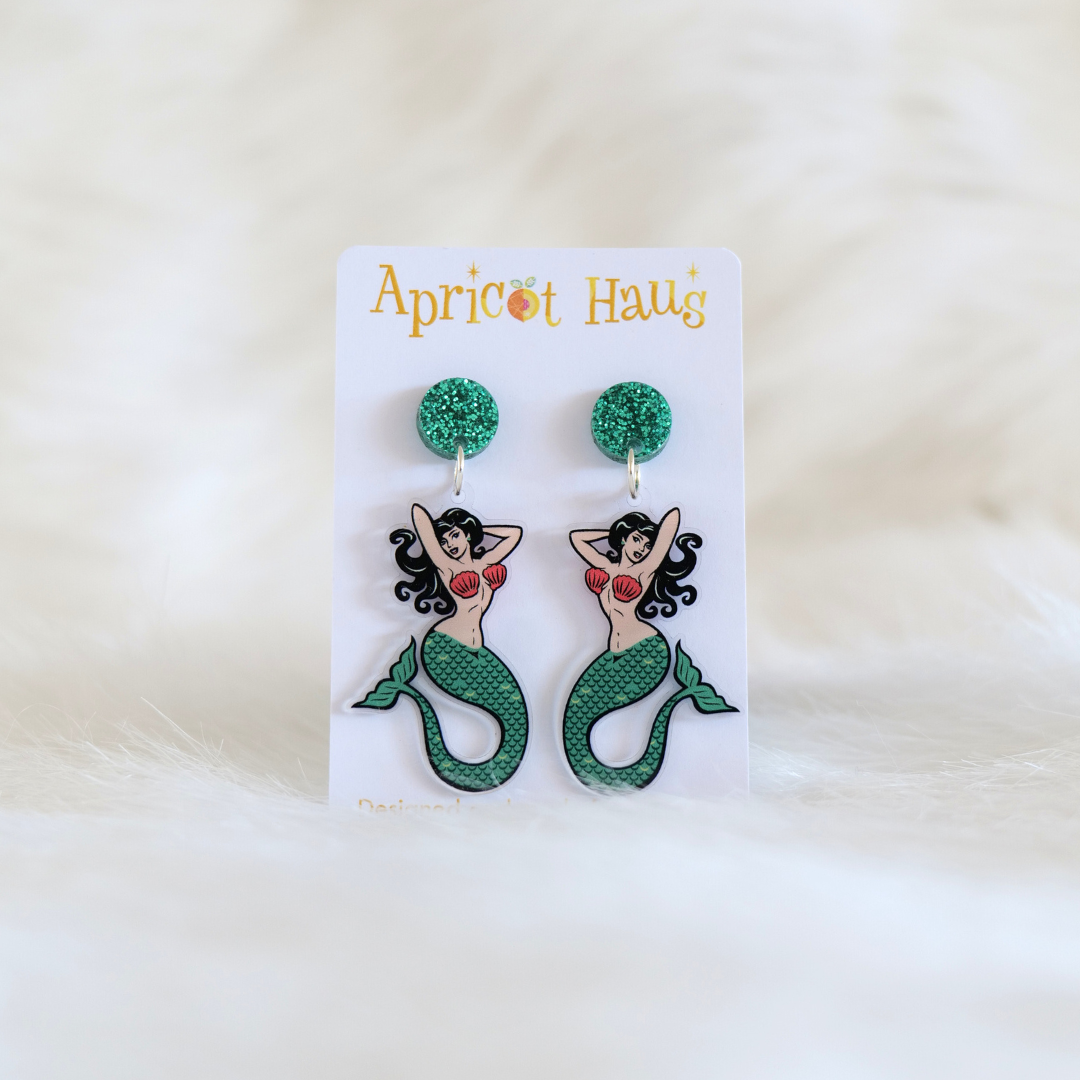 Pair of acrylic earrings in retro tatto-style mermaids with pink clam shell bikini tops and teal tails and circular teal sparkly tops; displayed on an Apricot Haus earring card on a white background