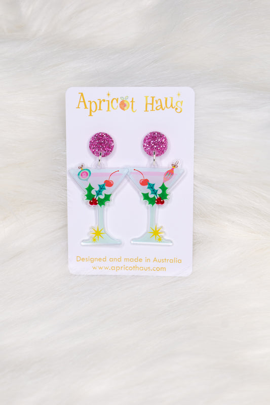 Earrings in the shape of martini glasses, decorated with holly, Christmas ornaments and a cherry, with a pink glitter stud at the top; displayed on an Apricot Haus earring card on a white background