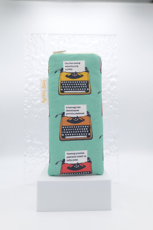 Glasses case featuring Pangram Typewriters design with illustrated vintage typewriters each typing a different pangram (sentences using every letter of the alphabet) on a soft green background; made from cotton canvas with a zip-top closure, padded and lined to protect lenses, handmade in Melbourne and certified Australian Made; pattern placement may vary slightly due to the handmade nature of the product.