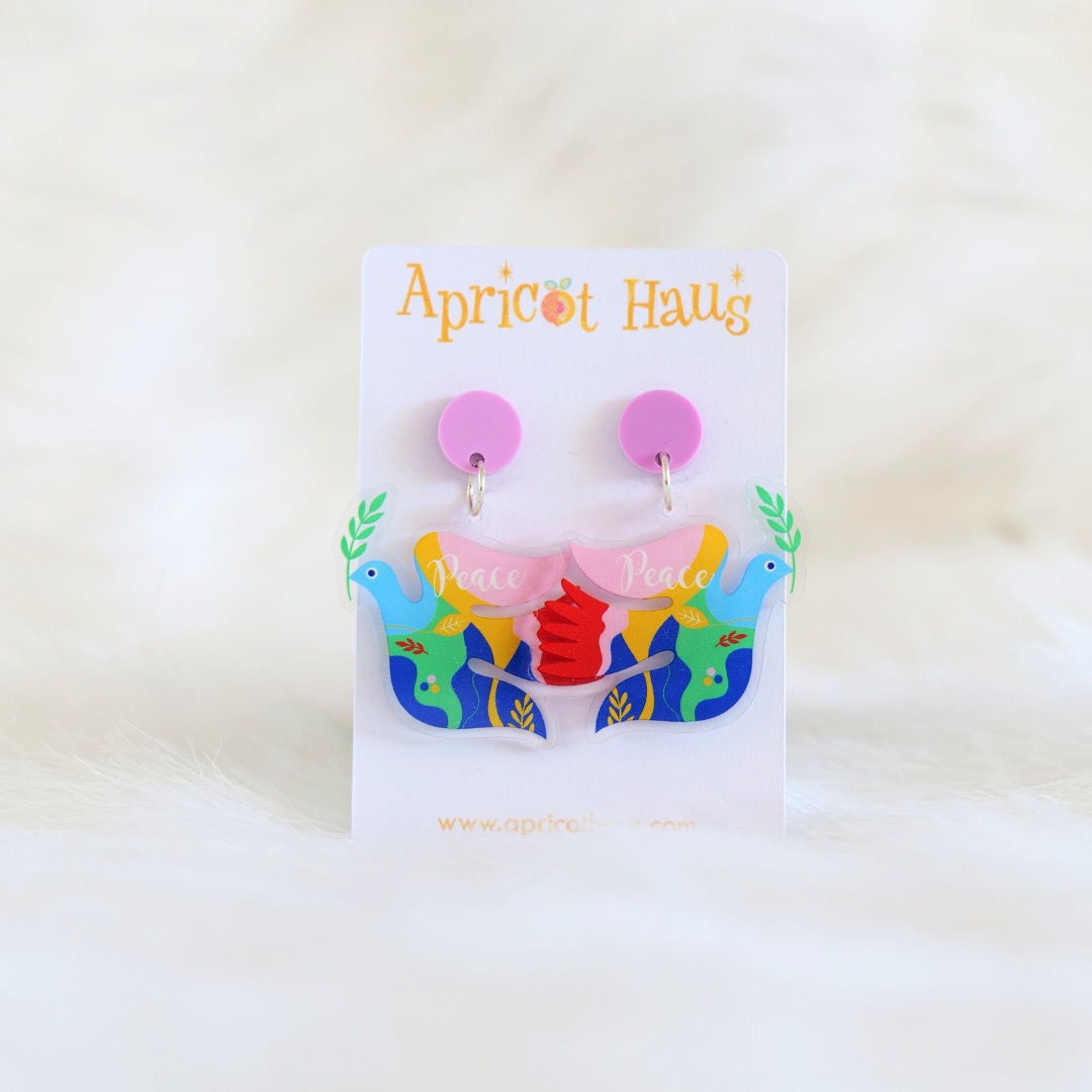 Pair of lightweight acrylic earrings in the shape of a dove with multicolour patches, a branch in the beak and the word peace across the top wing; earrings have a circular pink top and are displayed on an Apricot Haus earring card