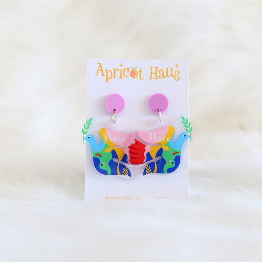 Pair of lightweight acrylic earrings in the shape of a dove with multicolour patches, a branch in the beak and the word peace across the top wing; earrings have a circular pink top and are displayed on an Apricot Haus earring card
