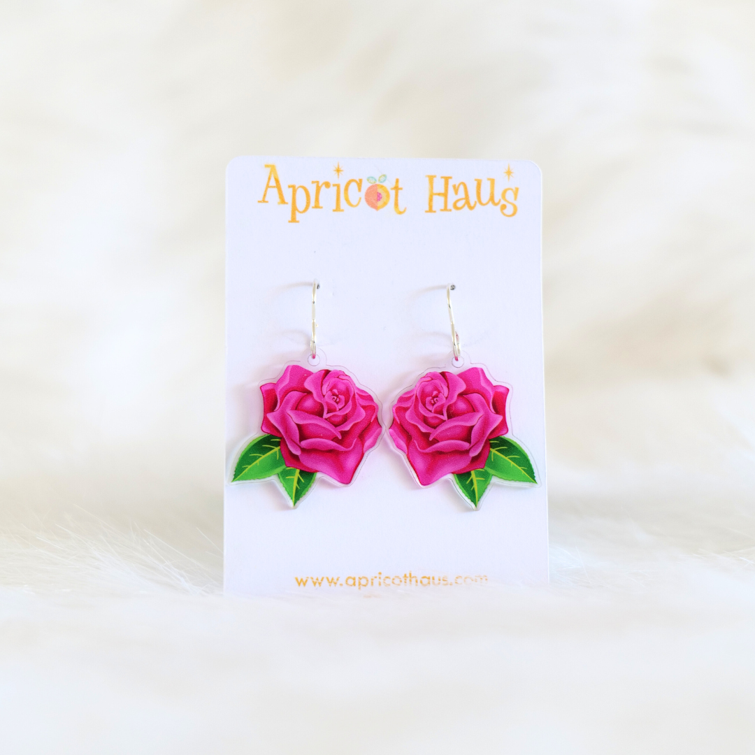Pair of acrylic earrings in the shape of a pink rose with two green leaves, lightweight with stainless steel hooks; designed and handmade in Australia, displayed on an Apricot Haus earring card