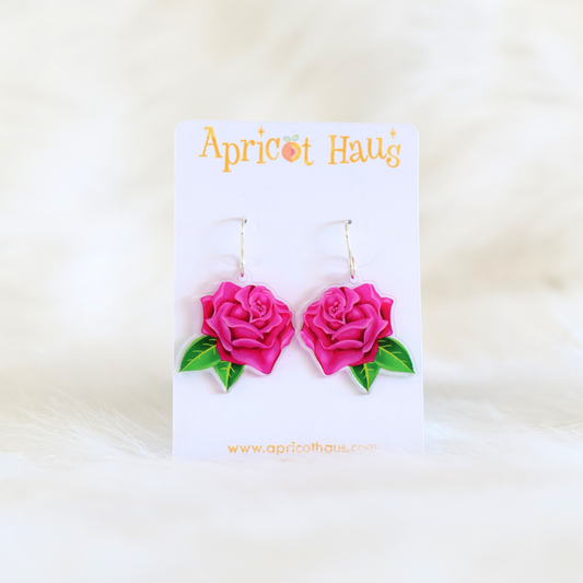 Pair of acrylic earrings in the shape of a pink rose with two green leaves, lightweight with stainless steel hooks; designed and handmade in Australia, displayed on an Apricot Haus earring card