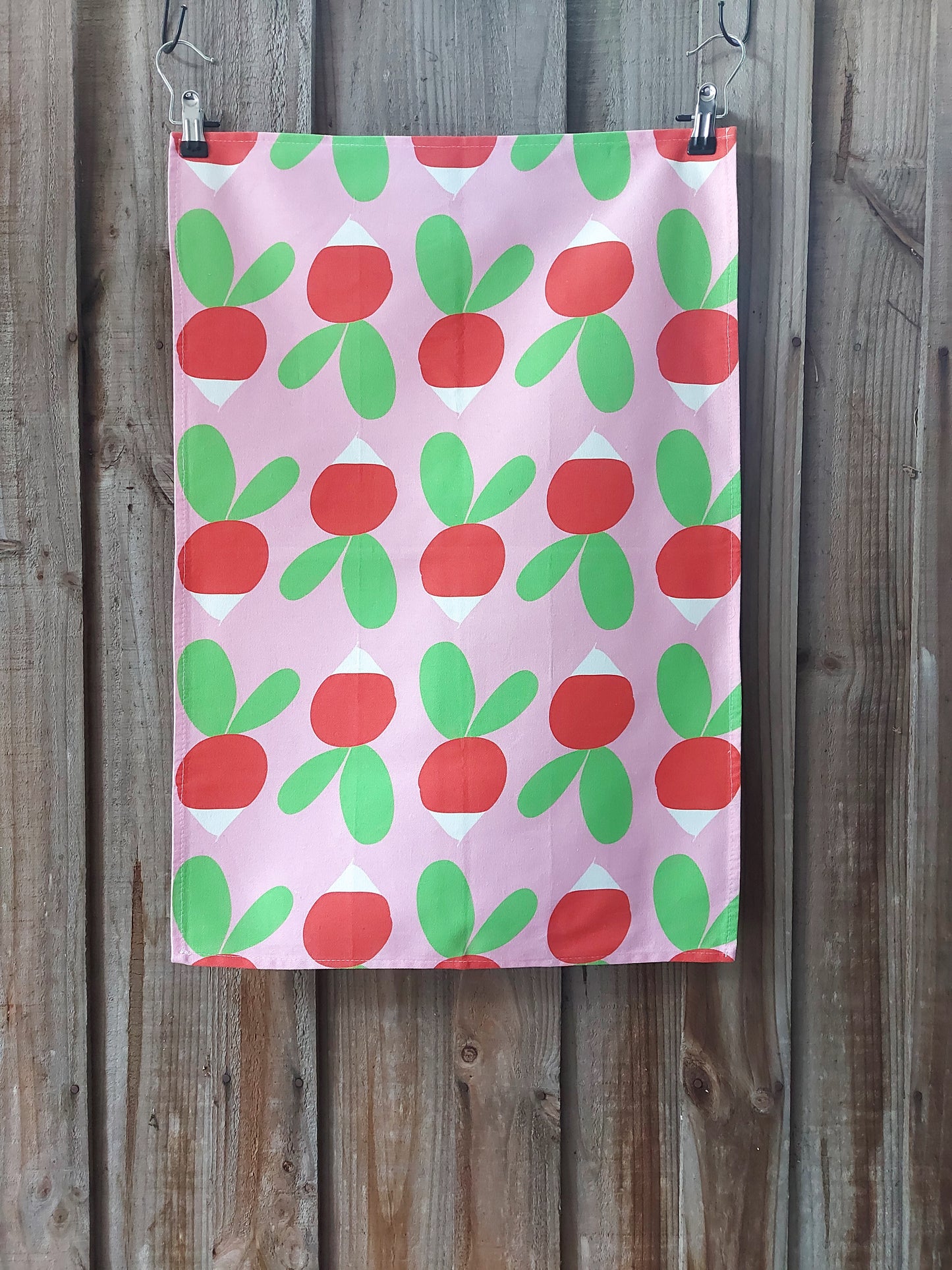 Cotton tea towel featuring bright red and green radishes in Scandinavian inspired design on a pink background; displayed against a wooden fence
