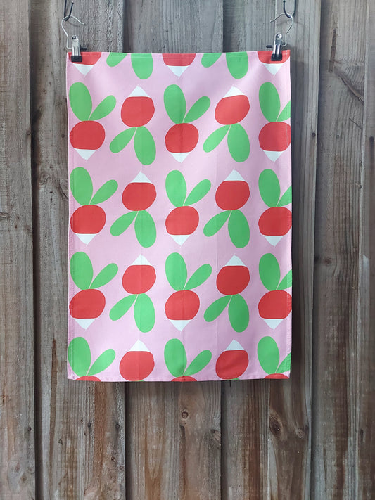 Cotton tea towel featuring bright red and green radishes in Scandinavian inspired design on a pink background; displayed against a wooden fence