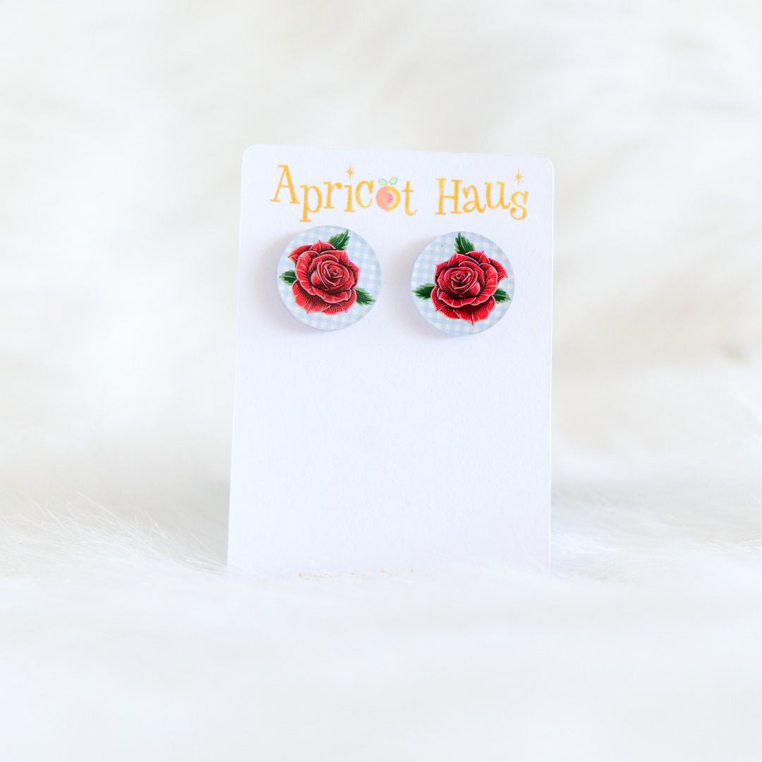 Two 2cm round stud earrings featuring a traditional style red rose on a soft blue and white check background, printed on acrylic with stainless steel posts and large accessible backs, displayed on an Apricot Haus earring card