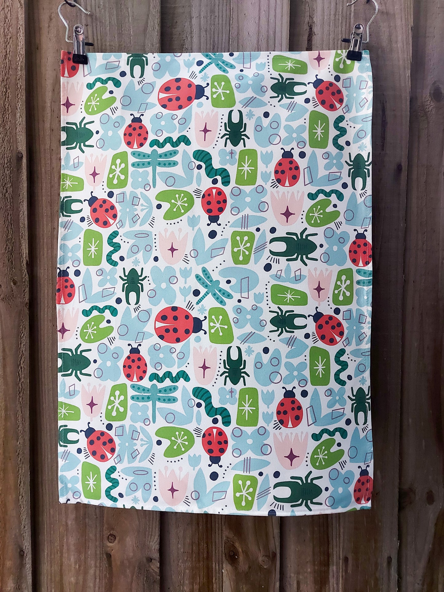 100% cotton tea towel featuring retro‑style illustrations of beetles, ladybirds, dragonflies and geometric garden motifs in a mid‑century colour palette; displayed against a wooden fence