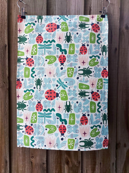 100% cotton tea towel featuring retro‑style illustrations of beetles, ladybirds, dragonflies and geometric garden motifs in a mid‑century colour palette; displayed against a wooden fence