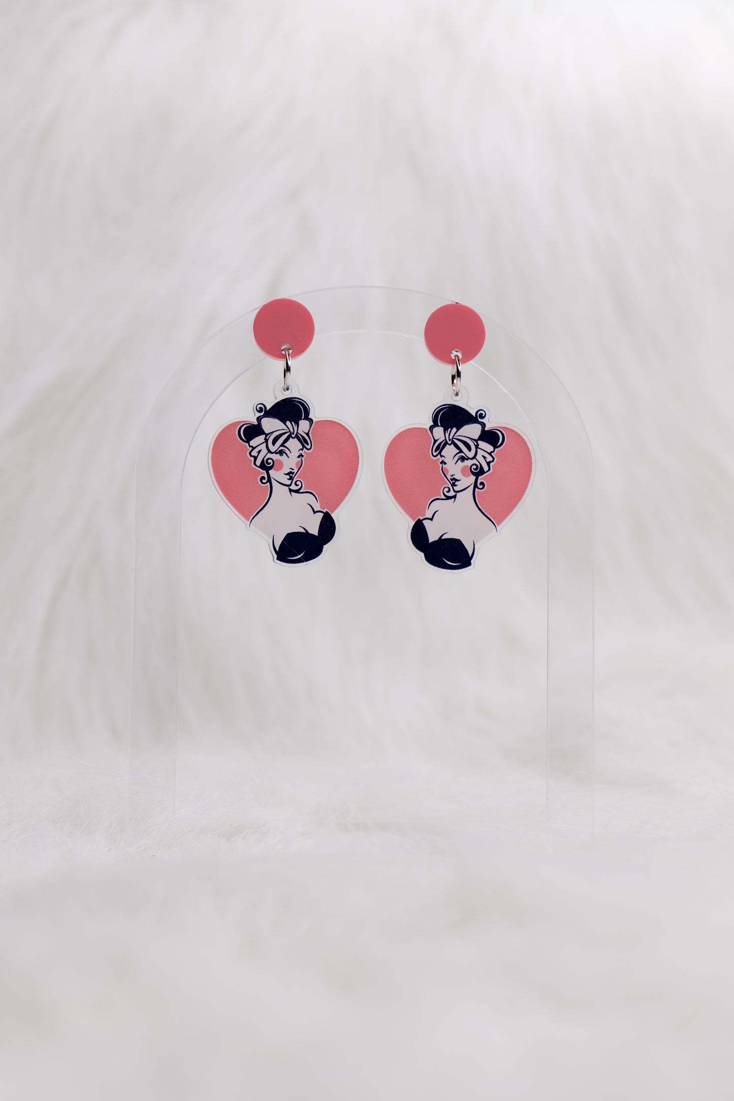 Pair of acrylic earrings with a pink circular stud and heart-shaped drops featuring a retro pin-up styled illustration of a girl with dark hair and bow; lightweight with stainless steel posts and large backs for accessibility, displayed against a white backgound 
